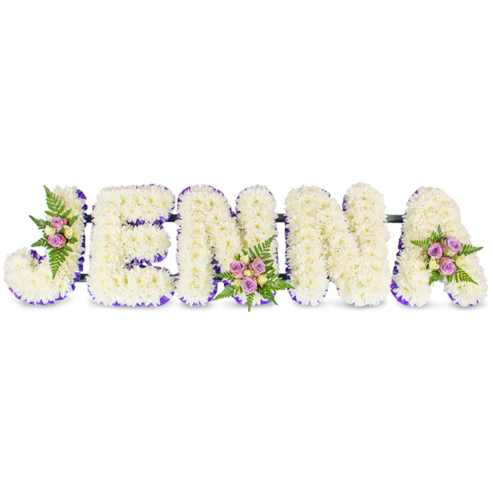 A lovingly crafted, personalised memorial arrangement spells out the name JENNA in substantial, three-dimensional capital letters, each composed of a dense carpet of creamy-white chrysanthemums or comparable small florets. The tight, cloud-like texture of the petals creates a soft, comforting surface that looks both refined and deeply respectful against the immaculate white background. Each letter is edged with a vivid stripe of deep purple - either a satin ribbon or a ring of tiny lavender blooms - which adds a clear visual boundary and a dignified accent. Three small, artfully placed floral clusters enhance the design: one nestles into the curve of the J, another rests at the base centre of the first N, and a third sits upon the upper arm of the A. These mini posies combine blush-pink and pale lavender spray roses with a scattering of cream or pale yellow roses, all cushioned by dark green fern fronds that introduce a fresh, leafy texture and a subtle herbal scent. Thin, dark structural bars connect the letters discreetly, indicating the kind of stable support used for chapel displays or cemetery tributes. The soft, even lighting highlights petal form and ribbon sheen, lending a serene, contemplative atmosphere. As Stanmore florists often advise, this understated style is suited to wakes, funerals, and memorial services across Stanmore Broadway, by Stanmore Tube and around Bentley Priory; Flower Delivery Stanmore can prepare this bespoke name tribute for local delivery and chapel placement with care and discretion.