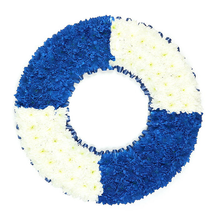A beautifully designed lifebuoy-style floral wreath dominates this image, resting against a stark white background that spotlights its intricate craftsmanship. The wreath forms a large, symmetrical circle with a prominent central opening, imitating the reassuring outline of a life ring seen on a ship's deck. Small, densely packed flowers, likely chrysanthemums or similar long-lasting blooms, cover the entire surface in a uniform layer, so that the ring appears lush and substantial. The colour arrangement is thoughtfully planned: four equal quadrants alternate between lush royal blue and clean, snow-white flowers. The top left and bottom right are filled with the velvety, saturated blue blooms, evoking the quiet depth of open water, while the top right and bottom left radiate with white blossoms that suggest clarity, remembrance, and peace. A delicate ribbon detail runs along the inner circumference, where a dark blue ribbon is neatly pleated to create a subtle frill, adding another dimension to the composition and reinforcing the maritime character. The overall look is both modern and respectful, ideal for a funeral tribute that needs to communicate strong emotional meaning without visual clutter. In Stanmore, a wreath like this might be chosen for a loved one who once navigated real seas or simply navigated life's challenges with calm resolve, perhaps someone remembered walking through Canons Park or along the leafy streets off The Broadway. It would sit gracefully by a coffin or memorial, conveying themes of safe harbour, loyal companionship, and a guiding presence that remains cherished even after farewell.