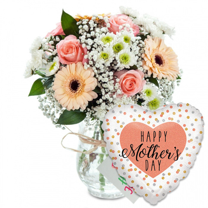 A serene Mother's Day scene is captured here against a plain white backdrop, allowing the soft colours and textures of the flowers to tell their own story, much like a quiet spring morning in Stanmore's residential streets. On the left, a rounded bouquet overflows from a clear glass vase shaped like a simple jar. Around the neck of the vase, a slim band of rustic twine is tied in a modest bow, lending the arrangement a handcrafted charm you might find at a small local florist near Stanmore Broadway. The bouquet itself is a celebration of pastel warmth. Clusters of peach and light pink roses are fully opened, revealing layers of delicate, spiralling petals that form gentle cups of colour. Among them, two or more striking peach gerbera daisies stretch their rounded petals outward, each bloom centred with a rich, dark brown eye that anchors the design. Weaving through these focal flowers, delicate sprays of white baby's breath soften every edge, while smaller white chrysanthemum blooms with fresh pale green centres nestle between, adding depth and fullness. Glossy green foliage supports and frames the blossoms, creating a natural backdrop reminiscent of the leafy paths leading towards Bentley Priory Nature Reserve. Just in front and slightly to the right, a heart-shaped foil balloon leans into the arrangement. Its smooth white surface is scattered with bright metallic gold and silver dots, catching imaginary light as if from a party in a Stanmore living room. At its centre, a large peach-coloured heart holds the words HAPPY Mother's DAY in bold black text; HAPPY is simple and clear, while Mother's Day is written in elegant, flowing script. Nestled quietly between vase and balloon is the corner of a small white greeting card, completing the sense of a carefully prepared, heartfelt gift.