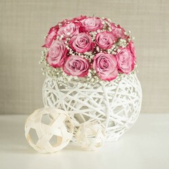 A detailed still life captures a romantic pink rose arrangement that could easily sit on a polished sideboard in a Stanmore townhouse, bringing softness to a calm, neutral room. At the heart of the image is a compact, dome-shaped bouquet of soft pink roses, about fifteen or more stems, each rose fully formed yet still slightly cupped, with petals that fade from a pale, almost milky blush in the centre to a more saturated, almost fuchsia pink along the ruffled outer edges. Threaded between the roses are generous clusters of white baby's breath, their miniature blooms forming a misty veil over the darker green stems, which gives the whole arrangement a light, airy feel. The flowers are set into a distinctive spherical vase made from white-painted woven fibres, arranged in an open lattice pattern like a modern reimagining of wicker. The structure of the vase creates soft pockets of shadow and light on the white tabletop beneath, suggesting gentle daylight filtering in, perhaps like the natural brightness that fills homes near Stanmore Country Park or along The Broadway. Beside the vase, two decorative orbs made from pale, interwoven wooden strips introduce an extra note of natural texture; one ball is larger and sits closer to the viewer, while the smaller one tucks slightly behind. The background is a warm, soft beige wall with a subtle texture that doesn't compete with the flowers, allowing the blush tones and crisp whites to stand out. The composition evokes quiet celebration and understated elegance, well suited to an intimate birthday surprise, a Mother's Day gesture, or a simple thank-you delivered to a friend in Stanmore.