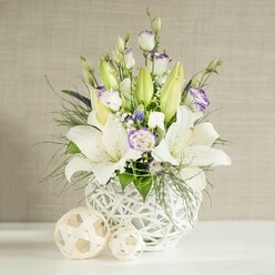 An elegant wedding-style centerpiece of lilies and lisianthus is captured in soft, even light, as if placed on a reception table in Stanmore, close to the greenery of Stanmore Country Park. The flowers emerge from a distinctive round white vase with an open, basket-like lattice that allows shadows and highlights to play across its surface, giving the design a sense of lightness. Majestic white lilies form the focal point, their trumpet-shaped blooms fully open, petals arching back to reveal pale green centres and delicately dusted stamens. Taller green lily buds push upwards within the arrangement, suggesting upcoming blooms and adding height and freshness. Around these central lilies, lisianthus blossoms in white and cream offer a softer texture, some petals traced with a gentle touch of lavender-purple along their ruffled edges. Tiny white filler flowers and a scattering of small, deeper purple accents are tucked between the larger blooms, enriching the bouquet with subtle detail. The foliage is abundant yet thoughtfully placed, with feathery, fern-like greens and broader leaves spilling lightly over the rim of the vase, creating an organic, slightly cascading shape that feels natural and welcoming. On the pristine white table beneath, two decorative spheres woven from a pale beige material rest casually at the side, echoing the vase's texture and tying the composition together. Behind, a light beige wall with a horizontal, linen-style texture forms a quiet backdrop, its understated pattern giving depth without distraction. The overall mood is calm, pure, and celebratory-a versatile floral design suitable for wedding tables, anniversaries, or heartfelt sympathy gestures delivered across Stanmore by local florists who understand the area's gentle elegance.