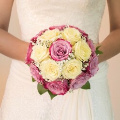Amid the serene beauty that defines Stanmore's historic lanes, a bride gently holds a vibrant, hand-tied bouquet-a statement of both love and artistry by local florists. The tightly woven arrangement is a visual celebration: creamy, sunlit yellow roses cluster together, cosseted by rich fuchsia roses and softer pinks touched with lavender, their petals overlapping in careful harmony. Wisps of delicate white gypsophila, like fairy-tale clouds, weave through the blooms, highlighting the bouquet's rounded form and creating a light, floating effect. The bouquet is encircled by rich, dark leaves providing contrast and lushness. The bride's wedding gown, with its ornate lace bodice and gracefully falling ivory skirt, beautifully frames the flowers, which she holds at her waist, radiating anticipation and quiet joy. Bright yet gentle light animates every detail, each petal's satiny sheen mirrored by the fabric of her dress. The softly blurred background, reminiscent of Stanmore's open greens and intimate garden spaces, ensures that the bouquet remains the emotional centerpiece-a symbol of devotion, refined taste, and the cherished local tradition of thoughtful, elegant floral design.