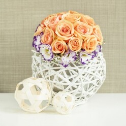 A beautifully balanced floral arrangement sits poised on a spotless white tabletop, as if ready to grace a wedding table in a Stanmore venue overlooking the greenery of Canons Park. The design is centred around a generous dome of peach and apricot roses, gathered so tightly that their blooms create a satisfyingly smooth, rounded form. Each rose is fully open, its petals thick and velvety, curving back just enough to reveal intricate spirals at the centre and subtle shifts of colour from soft blush to richer apricot tones. At the base of this rose sphere, a delicate ring of lisianthus blooms wraps around like a collar. Their white petals are beautifully edged with deep violet and purple, adding a refined contrast that feels both dramatic and gentle at once. Interspersed throughout are fine sprigs of white baby's breath, their clusters of tiny blossoms forming a lacy, cloud-like layer that softens the transitions between roses and lisianthus. The entire composition is anchored in a distinctive white woven container, broadly spherical and slightly bulbous, made from interlaced strands of wicker or rattan. Its open, lattice-work construction lets the light filter through, casting faint shadows on the table and adding a sense of lightness despite its generous size. In front of the arrangement rest two decorative spheres woven from pale, natural wood-toned strips, one noticeably larger than the other. Their open, airy structure complements the vase, lending the scene an artisanal touch that would suit the calm elegance of Stanmore's family homes. The soft, subtly textured beige background completes the picture, creating a warm, neutral setting that highlights the fresh blooms and makes this composition perfect for special occasions, refined home décor, or wedding styling across Stanmore.
