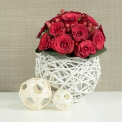 A rich, romantic centerpiece of red roses is showcased against a clean, neutral backdrop, as though prepared for a sophisticated celebration in a Stanmore home. At the heart of the scene sits a perfectly rounded bouquet of crimson roses, closely clustered together to form a smooth dome. Each rose is caught at a slightly different moment of its bloom, from tightly spiraled buds to more open blossoms, their velvety petals creating a plush, tactile surface that invites the imagination to trace each curl. Small clusters of glossy red hypericum berries are carefully placed among the roses, their polished, bead-like forms introducing a playful contrast and a lighter sheen of red, reminiscent of fruiting shrubs in gardens around Bentley Priory. Just beneath the mass of blooms, a discreet layer of dark green foliage offers quiet support and a muted frame for the vivid colors above. The bouquet is nestled into a distinctive spherical vase made from white-painted wicker or twigs, woven in an open, cage-like pattern. This airy globe allows threads of light to pass through, casting delicate shadows on the smooth white tabletop and adding a contemporary twist to the rustic material. To the left of the main arrangement, two decorative wicker balls in natural beige tones lie on the same surface. One is larger and more prominent, while the smaller sphere tucks in behind it; both echo the vase's interwoven texture in softer, natural wood hues that would feel at home in an elegant Stanmore dining room near Stanmore Hill. Behind the arrangement, a softly blurred beige or taupe wall, with a subtle woven or plaster-like finish, recedes gently into the background. The overall mood is intimate, polished, and quietly celebratory, ideal for wedding receptions, anniversaries, or special dinners, with flowers delivered fresh across Stanmore.
