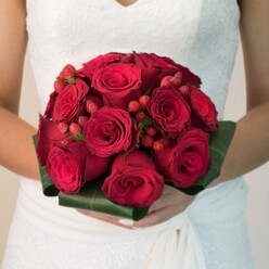 Bathed in soft, early afternoon light perhaps just outside Stanmore Hall, this image draws the eye to a bride's hands gracefully holding a spectacular, rounded bouquet crafted for both impact and intimacy. Each velvety red rose, in bloom or budding, is artfully nestled among rich, glossy greenery that forms a sculpted collar-a hallmark of classic wedding floristry. Tucked between the scarlet blossoms are clusters of vibrant red hypericum berries, their doll-like orbs offering a playful visual counterpoint and a hint of local garden abundance. The overlapping petals and berries create a sense of layered depth and tactile invitation, the bouquet's perfume subtly described as a blend of sweet rose and the earthiness of Stanmore's leafy parks. The bride wears a snow-white dress with intricate lacework along the bodice and a soft chiffon skirt that brushes gently out of frame, a vision reminiscent of timeless vows exchanged at St. John's Church. There's a rhythmic unity in the composition-abundant yet perfectly balanced-mirroring both the joy and the serenity of a memorable wedding morning in Stanmore. The floral artistry here isn't just decorative; it encapsulates cherished tradition and the vibrant promise of new beginnings within a beloved North London community.