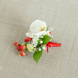 A refined floral buttonhole lies neatly centred on a natural, light beige woven cloth, the subtle texture of the fabric echoing the rustic elegance often chosen for weddings and civil ceremonies around Stanmore Country Park. The eye is immediately drawn to a flawless white Phalaenopsis orchid, its velvety petals fanning open in a calm, symmetrical curve, while the gently ruffled throat reveals a blend of creamy yellow and soft orange, like a quiet glow at the heart of the flower. Encircling this central bloom, airy sprays of baby's breath form tiny constellations of white, each miniature blossom contributing to a soft, mist-like halo that lightens the composition. Nestled alongside them is a compact cluster of high-gloss red berries, round and full, their deep cherry tones adding a joyful, almost festive accent, complemented by a few paler green berry buds that bring a sense of freshness and new growth. Polished green leaves, some broad and pointed, others slimmer and more elongated, are carefully placed to cradle the orchid and berries, wrapping the piece in gentle greenery and giving it a well-balanced silhouette against the neutral background. At the base, the florist has bound the stems in a vivid red satin ribbon, smooth and slightly reflective, tied into a tidy knot so that the short ends rest gracefully along the cloth, ready to be fastened to a jacket. The image captures not just the colours and textures, but the feeling of poised anticipation, as though this boutonniere is waiting for the moment it will accompany its wearer to a special celebration in or around central Stanmore.