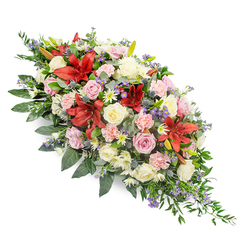 This memorial spray stretches diagonally across a pure white backdrop, its colours blooming like a soft, steady breath of comfort. Crafted in a style often requested by families in Stanmore and along The Broadway, the piece feels both traditional and thoughtfully modern. Generous deep red lilies, their star-shaped petals fully unfurled, provide powerful focal points, radiating warmth and strength at the centre of the design. Around these, clusters of creamy white roses and gentle blush and pale pink roses are arranged at different heights and stages of opening, their velvety petals layered like unfolding memories. Between them, light pink carnations with delicately ruffled edges blend seamlessly into smaller white chrysanthemum-like blooms and daisy-inspired flowers, giving the spray its lush, abundant texture. Cooling the palette, spiky blue-purple thistles and clusters of tiny lavender-blue filler flowers are woven through the arrangement, adding depth, contrast, and a subtle suggestion of the wild, similar to the natural feel of Stanmore Country Park. A generous bed of dark green foliage cradles the entire display, with broad, glossy leaves providing structure and narrower, fern-like sprigs softening the perimeter. The greenery extends slightly beyond the blooms, particularly towards the tapered lower edge, creating a flowing outline that leads the eye gently along the piece. The overall mood is one of calm reflection and gradual healing, the kind of floral tribute that brings a sense of peace to a chapel or crematorium service. It speaks in quiet tones of love, remembrance, and the enduring impact of a life, making it a meaningful choice for a Stanmore family saying goodbye.