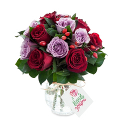 This arrangement reads like a quiet love letter rendered in flowers: a tight, round composition of deep crimson and gentle lavender roses, each bloom open and layered so their petals catch and hold the light. The reds vary from warm scarlet to almost-burgundy, the purples from soft lilac to dusty mauve, and together they form a slow, harmonious colour conversation across the bouquet. Interwoven dark green leaves - the kind of glossy Ruscus or Salal we use in the shop - provide cool relief and sculpt the overall form, while clusters of orange-red rose hips add small bright notes, like berries hiding in hedgerow after rain. The blooms sit in a bulbous, mercury-glass vase whose pitted silver surface reflects and softens highlights; natural raffia is knotted at the neck, lending a measured rusticity to the otherwise elegant design. A small white gift tag dangles forward, hand-angled to catch the eye and bearing a tiny floral sketch rather than a corporate mark, suggesting a personal message inside. Against a clean white backdrop the colours sing with clarity; the tactile contrasts - satiny petals, firm berry skins, smooth glass - are almost palpable. Presented as a thoughtful gesture, this bouquet suits anniversary tables, quiet celebrations, or bedside brightening, and it travels well for local delivery across Stanmore and nearby streets such as Stanmore Broadway and Stanmore Hill when you want to give the impression of considered, artisan care.
