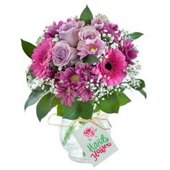 This sumptuous bouquet, crafted by a skilled Stanmore florist, tells a quiet tale of affection and joy-each element thoughtfully chosen to convey grace and warmth. Elegant lavender roses take center stage, their petals gently curling outward as if coaxed open by the peaceful morning air drifting over Stanmore Hill. Around these, striking Gerbera daisies bloom in hot fuchsia, their bold faces and velvety dark cores exuding playful energy. Filling the arrangement, magenta chrysanthemums lend depth and complexity, their rich hues reminiscent of the vibrant colours seen at local gardens after spring rain. Sprigs of baby's breath add a delicate, airy halo, while deep green leaves around the base introduce movement and a touch of wildness. This lush ensemble is arranged in a crystal-clear vase, its unique cracked-ice texture catching the sunlight and adding character. A band of natural raffia is tied neatly at the neck, holding a blank, white gift tag, ready for messages of joy or heartfelt wishes. The bouquet's full, abundant shape and gentle colour harmonies make it a perfect choice for everything from milestone birthdays to simply sending an uplifting surprise to friends in Stanmore, carrying a whisper of soft perfume and the promise of freshness with every delivery.
