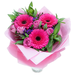 This arrangement reads like a springtime promise, carefully balanced between exuberance and tenderness. Three vivid Gerbera daisies in a saturated fuchsia take centre stage, each bloom opening wide to reveal a dark, grounding centre that gives the bouquet a striking focal point. Interspersed are roses in a muted lavender-pink-fresh, tightly budded forms whose velvet petals catch the light and lend a romantic softness. Little clusters of purple statice weave through the composition, their fine texture and cooler tone creating playful punctuation among the larger blooms. A backdrop of lush green foliage-broad, glossy leaves reminiscent of salal or eucalyptus-adds volume and a clean, botanical freshness. The bouquet is wrapped with intention: the inner matte pale-pink tissue cushions the stems while a translucent, slightly crinkled fuchsia-pink cellophane wraps around the blooms and introduces a gentle shimmer that unifies the colour palette. The lower edge of a clear glass vase is visible, assuring the recipient that the flowers are fresh and display-ready. In morning light the colours glow warmly; the roses give off a soft, floral scent while the greenery offers an aromatic, green counterpoint. Ideal for celebrating birthdays, anniversaries, Mother's Day, or for a cheerful surprise, this hand-tied composition is the kind of piece a customer might order for same-day delivery around Stanmore Station, Stanmore Broadway, or the surrounding neighbourhoods. It's intimate, bright, and made to make someone feel remembered.