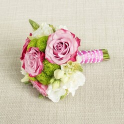 Resting on a pale, sand-toned fabric that recalls a linen table runner in a Stanmore townhouse, this compact bouquet captures a poised moment of celebration. Three pink roses form the heart of the design, two of them fully open with luxurious, spiralling petals, and a third slightly more modest yet equally graceful. Their colour drifts from soft pastel pink at the centre to richer, magenta-tinted edges, giving each bloom a gently ruffled, painterly look. Between the roses, vibrant lime-green button chrysanthemums form rounded clusters, their countless tiny petals creating a cushion-like texture that almost invites touch. Threaded through this interplay of pink and green are airy white and cream blossoms, likely lisianthus or freesia, their slender petals curling outward like whispers of silk. Small, star-shaped white flowers with yellow centres punctuate the arrangement with delicate points of brightness, reminiscent of hedgerow blossoms seen on a walk through Canons Park. The stems, cut short and tidy, are firmly wrapped in vivid fuchsia satin ribbon, its surface smooth and gleaming in the soft light. Over the ribbon, a slim strand of white faux pearls winds diagonally, adding a note of vintage romance, as if chosen to echo bridal jewellery. The lightly textured background, with its subtle weave, acts as a calm, neutral stage for the bouquet's colours to glow. This intimate arrangement feels perfect for a bridesmaid's bouquet at a Stanmore registry ceremony, a chic engagement celebration at home, or a sweet gift delivered locally, designed as much for the hand that cradles it as for the eyes that admire it.