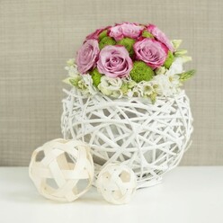 A stylish floral centerpiece, perfect for a wedding in Stanmore or a special family gathering near Stanmore Hill, is showcased in this image. At the heart of the composition is a compact, domed bouquet brimming with roses in shades of pink and lavender. The roses are in full bloom, their petals opening into generous spirals, with some edged in a more intense pink that creates a gentle ombré effect. Among the roses, bright green button chrysanthemums provide crisp bursts of colour and structure, their rounded forms and densely textured petals adding a playful, contemporary touch. Around the base of the main bouquet, delicate white filler flowers weave in and out, softening the outline and giving the impression that the blooms are subtly overflowing from their container. The arrangement is held in a spherical white vase made from slender, rattan-like strands woven together in an open lattice design, leaving airy gaps that allow light and shadow to interplay. This vessel has a modern rustic charm, like something you might discover in a boutique home store near Stanmore Broadway. To the left on the same clean white surface, two decorative spheres made from pale wicker or twigs, one larger and one smaller, mirror the vase's weave and introduce an extra layer of texture and interest. The background is a softly defocused beige fabric, its gentle texture reminiscent of natural linen, providing a neutral backdrop that allows the pinks, lavenders, and greens of the flowers to stand out vividly. The overall effect is one of understated luxury and warmth, ideal as a wedding table arrangement, centerpiece for a bridal shower, or an elegant accent in a Stanmore home.