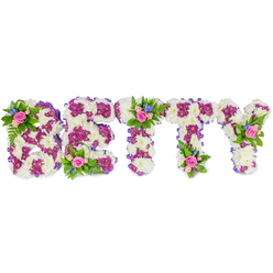 This bespoke funeral tribute spells BETTY in substantial, freestanding capital letters, each letter lovingly formed from a dense base of creamy-white chrysanthemums that give a plush, cloud-like texture beneath soft overhead lighting. Deep magenta and purple chrysanthemums are used to carve and accentuate inner shapes and outlines, their jewel-toned clusters creating depth and a quiet vibrancy that reads with clarity from a distance. Along the edges, a slim strip of purple ribbon tucks in like a finishing seam, lending cohesion and a subtle touch of formality. Small clustered bouquets crown the B, the central cross of the first T, and the upper arm of the Y; these accents contain blush-pink roses in mixed stages of bloom, small bright blue filler flowers that could be delphiniums or cornflowers, and delicate fern fronds that trail slightly, softening the geometry. The pink roses offer a gentle, familiar fragrance while the blue accents bring a crisp visual counterpoint; together the palette feels composed, sympathetic and quietly hopeful. Presented against a clean white background, the tribute's textures-from the chrysanthemum's cushiony heads to the satin of ribbon and the fern's crisp fronds-are clearly visible and tactile in description. As a Stanmore florist who has arranged memorial pieces for families around Stanmore Station and Stanmore Hill, Flower Delivery Stanmore prepares tributes like this with the care and restraint appropriate for a wake or celebration of life, intended to honour memory with handcrafted detail and local sensitivity.