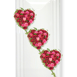 Three luxurious hearts made entirely of roses form the focus of this moving funeral tribute, each heart stacked beneath the other in a graceful diagonal, as if one memory flows into the next. The hearts are full and robust, shaped by hundreds of tightly nestled roses in deep crimson red, bright fuchsia, and delicate blush pink. The darkest red blooms give the piece a sense of solemnity and unwavering devotion, while the pink tones introduce softness, compassion, and affectionate recollection. Petals layer over petals so that the hearts take on a gently rounded, almost cushion-like look, inviting the viewer to imagine their velvety texture and faint, sweet fragrance. Framing each heart, slim tendrils of fresh ivy trace the curves in a continuous line of glossy green, then trail downwards to link to the heart below, creating a visual metaphor for the threads of love and family that continue long after a goodbye. The design sits against a pure white background, similar to a calm, light-filled wall in a Stanmore memorial hall or a quiet room near Stanmore Country Park, where nothing distracts from the beauty of the flowers. Subtle shadows underline the three-dimensional shape of the hearts, making them feel almost suspended in front of the surface. This image captures the essence of a tribute that might grace a coffin, stand at the front of a service, or be placed beside a photograph of the departed, expressing in petals what may be difficult to say aloud: gratitude, devotion, and a bond that time cannot erase.