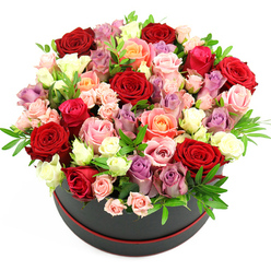 In this image, a luxurious round hatbox arrangement dominates the frame, isolated against a clean white backdrop that makes the colours appear especially vivid. The container is a sleek, matte black cylinder with a fine red line circling its base, a subtle yet stylish detail reminiscent of a designer gift box arriving at a Stanmore doorstep near The Broadway. Rising from the box is a densely packed, dome-shaped bouquet of roses, so full that the blooms billow above and beyond the rim. Deep red roses form the dramatic anchor of the composition, their velvety petals tightly layered and richly coloured. Interlaced with them are pale lavender, blush pink, and light fuchsia roses, each adding softness and romance. Warm peach and coral roses glow like little pockets of sunshine, while creamy white and ivory blossoms catch and reflect the light, lifting the entire palette. The arrangement includes both large, fully open roses showing off their spirals of petals and smaller, partially opened buds, as well as spray roses that introduce finer detail and rhythm. Tucked between the roses are slender stems of greenery, possibly eucalyptus or ruscus, their fresh green leaves offering a cool contrast and a hint of natural movement. The lighting is bright and even, making every curve, colour, and texture readily visible. The overall mood is modern yet timelessly romantic, ideal for marking anniversaries, engagements, milestone birthdays, or heartfelt thank-you moments around Stanmore. Each stem seems thoughtfully placed, reflecting the practiced eye of a local florist experienced in crafting statement hatbox designs that feel both luxurious and personal.