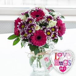 This tender, celebratory scene shows a carefully crafted bouquet and a cheerful heart-shaped balloon, presented together as if arranged by an experienced florist in Stanmore who knows exactly how to brighten someone's day. The flowers are displayed in a classic, rounded clear glass vase, filled with clean water that catches the light and reveals the straight green stems gathered neatly inside. Natural twine is tied around the neck of the vase in a relaxed bow, from which a small white tag dangles, printed with a stylised pink rose and a line of green script that looks like a friendly hand-written greeting, adding to the sense of a personalised gift. Rising from the vase, the bouquet forms a generous dome of colour. Large gerbera daisies in deep fuchsia are the first blooms to catch the eye, their smooth, radiating petals surrounding intense dark centres, giving the arrangement a bold, contemporary flair. Nestled among them are clusters of bi-coloured chrysanthemums, their petals a rich purple that fades to white at the tips, gathered around warm yellow centres that sparkle like tiny suns. Several creamy white roses are tucked throughout, their softly swirling petals lending elegance and a romantic softness. Fine sprays of white baby's breath drift between the bigger flowers, adding a delicate, cloud-like texture, while fresh green foliage frames and fills the bouquet, echoing the greenery of local spots like Bentley Priory Nature Reserve. To the right of the vase, a shiny foil balloon in the shape of a heart gleams under the light. It has a bright silver border and a white centre filled with playful, patterned letters in pink, purple, blue, and red that spell out "Love n' Hugs 24-7," complete with tiny hearts scattered around the words. Resting on a pristine white surface against a softly blurred, luminous background, this arrangement feels perfect for a same-day flower delivery in Stanmore when you want your message of love and hugs to last around the clock.