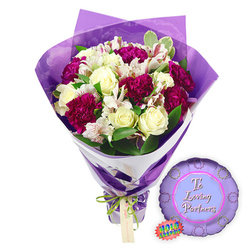 A carefully crafted bouquet and coordinating balloon sit on a plain white background, creating a sense of poised romance that would feel at home in any Stanmore living room overlooking the green spaces near Stanmore Country Park. At the centre of the bouquet are creamy white roses, each one tightly furled, the petals arranged in smooth spirals that suggest both freshness and enduring affection. Cradling them are generous heads of deep magenta and burgundy carnations, their softly frilled petals forming rich layers of texture and colour that bring warmth and intensity to the arrangement. Interwoven among the larger blooms are stems of white alstroemeria, their slender petals opened into trumpet shapes, delicately marked with reddish-brown speckles and fine stripes on the inner petals, offering an intricate, almost lace-like detail. Surrounding this mix, lush green foliage and touches of variegated leaves with pale edges provide contrast, fullness, and a hint of garden charm. 

The bouquet is wrapped in a refined two-layer presentation. Closest to the stems, a band of crisp white paper runs at a slight diagonal, giving the base a structured, contemporary look while supporting the flowers. Over this, a sheer purple cellophane wrapper rises above the blooms, lightly crinkled to catch the light and form a softly shimmering frame that echoes the tones of the carnations without overwhelming the whites. At the gathered stems, a thin, bright green satin ribbon is tied in a precise bow, from which hangs a small rectangular light brown gift tag, its blank surface suggesting a handwritten message yet to be added. To the right of the bouquet, placed on the white surface, rests a metallic purple foil balloon with a scalloped, rosette-like border. Its centre is a paler lavender disc, bearing the words "To Loving Partners" in elegant pink script, and a small jagged-edged holographic rainbow sticker near the lower left adds a playful flash of colour. This floral and balloon set offers a complete romantic gesture, ideal for anniversaries, proposals, or simply reminding a partner of your love anywhere in Stanmore or along the nearby Uxbridge Road.