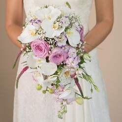 Within the serene atmosphere of a Stanmore morning, a bride is pictured holding a showstopping cascading bouquet, its effortless flow and exquisite layering drawing in the eye. The bouquet's shape mimics a waterfall, with radiant Phalaenopsis orchids at its core, their creamy white petals and gilded throats shining with natural grace. Surrounding them, blush pink and lavender roses bring classic refinement, some unfurling luxuriously while others remain as tightly closed buds-each symbolizing love's promise. The bouquet is lightened by feathery sprays of baby's breath, reminiscent of the gentle mists over Stanmore's green spaces at dawn. Touches of lavender and purple from small blossoms, perhaps sweet pea or stock, add depth, while vivid magenta trails of Amaranthus sweep downward, crafting movement and texture that energizes the design. Lush greenery and a few slender, unopened buds appear throughout, grounding the composition with a fresh, local touch. The bride's gown, its delicate lace overlay visible above the florals, contributes a sense of romance and local tradition. Set against a soft cream backdrop, the image radiates both poise and joy, much like a walk through Canons Park in early summer. This bouquet, created with expertise in Stanmore, offers a harmonious blend of modern flair and classic beauty-perfect for couples seeking floral artistry for their special day.