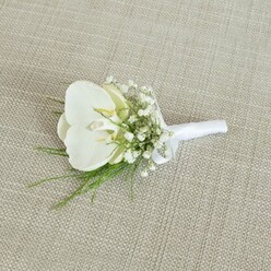 A refined white orchid boutonniere rests on a light tan, woven fabric surface that resembles natural linen, its subtle texture giving the image a warm, tactile quality. At the centre of this handcrafted piece is a flawless Phalaenopsis orchid, its creamy white petals gently curving outward like smooth porcelain, with a muted hint of pale yellow and soft ivory in the throat that suggests both freshness and gentle sweetness. Nestled around the orchid, airy tufts of white baby's breath form a delicate collar, each miniature flower catching the light and creating an almost misty, ethereal frame. Threading through this cloud of white are fine green stems and fronds of feathery foliage, adding structure, movement, and a whisper of woodland charm, as if freshly gathered from a sheltered garden in Stanmore. The lower portion of the boutonniere is meticulously bound in a bright white satin ribbon, wrapped tight and even, with a neat little bow positioned just where the stems begin, indicating it is ready to be pinned securely to a suit or tuxedo. The soft, neutral backdrop draws attention to the crisp contrast between the pure whites and lively greens, evoking the calm, anticipatory moments before a wedding ceremony in a local Stanmore venue or a stylish civil celebration nearby. This quietly luxurious buttonhole, the sort often prepared with care by a local florist at Flower Delivery Stanmore, captures the essence of modern elegance: simple lines, high-quality blooms, and an understated grace perfectly suited to grooms, groomsmen, or a distinguished guest at an intimate North West London celebration.