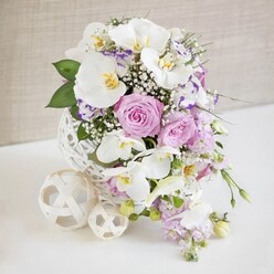 A beautifully composed floral arrangement sits centre stage on a white tabletop, as if ready to adorn a wedding table in Stanmore with understated luxury. The flowers are gathered in a distinctive round vase formed from white, interlaced strands that create a hollow, woven sphere, reminiscent of artistically crafted rattan. From this sculptural base, a rich cluster of white, pink, and purple flowers rises in a gentle dome, overflowing slightly at the sides. Several impressive white Phalaenopsis orchids stretch outward, their broad, smooth petals arranged like delicate fans, glowing softly under bright, even lighting. At the heart of each orchid, subtle yellow and golden tones provide a warm highlight, giving the design an exotic, polished character. Between the orchids, velvety roses in pastel pink and muted lavender nestle closely together, some spiralled tightly, others fully open to reveal lush, ruffled centres, echoing romantic celebrations and family gatherings held in Stanmore's elegant homes and venues. Smaller blossoms, likely Lisianthus, show white petals finely edged in purple or magenta, adding intricate detailing and colour depth. Threads of airy white baby's breath weave in and around the main flowers, creating a veil of tiny blooms that softens the composition. From one side, slender stems of pale lilac trailing flowers cascade gently downward, lending movement and a graceful, natural flow. Glossy green foliage peeks through the pastel blooms, giving structure and a fresh contrast. Resting on the pristine white surface next to the vase, two cream-toned woven spheres-one larger, one smaller-mirror the vase's texture and complete the arrangement. The scene is set against a neutral, lightly textured beige background that fades softly out of focus, allowing the colours and textures to stand forward and creating a serene, refined mood that suits weddings and special occasions across Stanmore.