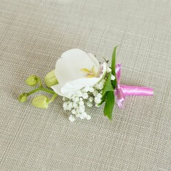 A single white orchid boutonniere is laid carefully across a pale beige, linen-textured fabric, its quiet elegance evoking the stillness just before a ceremony begins in one of Stanmore's churches or registry offices. The orchid, a Phalaenopsis bloom of pristine, milky white, forms the heart of the design, its broad petals smooth and slightly luminous, curving back to reveal a softly patterned centre with notes of buttery yellow and a whisper of pale pink. Encircling the base of this focal flower, wisps of white baby's breath gather in delicate clusters, their tiny rounded blossoms like a fine mist, adding a dreamy, ethereal quality that lightens the whole composition. A few narrow, rich green leaves peek out from behind, lending a fresh garden contrast and a hint of movement. Extending gently to one side, a slim stem carries a pair of tightly closed orchid buds in soft lime-green, their firm, satin-like surfaces suggesting life poised on the edge of blooming, much like the anticipation felt before vows are exchanged. At the base, the stems are bound with a narrow band of bright fuchsia satin ribbon, tied into a neat, tailored bow whose glossy finish shines subtly against the textured cloth. The pop of vivid pink feels joyful and modern, while the orchid and baby's breath keep the design classic and refined. This is the sort of handcrafted floral detail a local Stanmore florist might prepare for a groom, best man, or prom guest heading to a celebration near Canons Park or along Marsh Lane, adding a poetic yet polished finishing touch to their outfit.