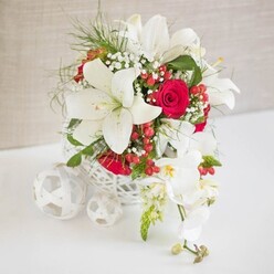 A luxurious floral design sits at the centre of a smooth white surface, bathed in soft, even light that brings out every nuance, as if styled for an intimate Stanmore wedding breakfast near Canons Park. The backdrop is a gently textured light beige fabric, creating a calm, neutral setting that allows the colours and shapes of the flowers to stand out. The arrangement itself is held in a round white container crafted with an intricate open-weave pattern, reminiscent of delicate wickerwork and adding a subtle, artisanal feel. From this sculptural base, several white oriental lilies rise and unfold, their elongated petals forming wide, elegant blooms around central stamens tipped with warm pollen. Tucked between the lilies are plush red roses, their velvety petals overlapping in tight spirals, deep and saturated in tone. Airy sprigs of white baby's breath weave between the larger flowers, lending a cloud-like softness and a touch of nostalgia, while slender stems of bright green foliage add structure, height, and a sense of movement, as if softly swaying in a Stanmore garden breeze. Glossy red hypericum berries appear at intervals throughout the design, offering small, round, jewel-like moments of colour that enhance the festive, celebratory feel. To the right, an elegant spray of white phalaenopsis orchids spills in a gentle cascade, each bloom pristine and slightly translucent, with delicate yellow and blush-pink touches at the centres, followed by a few tightly closed buds. On the left of the main bouquet, three white woven spheres rest on the tabletop, echoing the container's pattern and acting as modern decorative accents. The overall impression is of thoughtful craftsmanship and romantic elegance, making this arrangement perfect for wedding tables, anniversary dinners, or special celebrations in and around Stanmore.