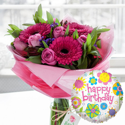A lush birthday bouquet and balloon gift set stands centre stage, as if waiting to be delivered across Stanmore by a local florist who knows just how to mark a special day. The arrangement is presented in a tall, clear glass vase, the stems neatly aligned and visible through the water, giving a sense of freshness and care. At the heart of the bouquet are large gerbera daisies in vivid shades of fuchsia and magenta, each bloom perfectly open with a dark, velvety central disc that anchors the composition. Wrapped around them are soft pink roses in varying moments of bloom, from tight buds with smooth, curved petals to partially opened flowers revealing swirls of delicate colour, suggesting both elegance and affection. Wisps of small, deep purple filler flowers weave between the larger heads, adding a hint of wildflower charm, while layers of fresh green foliage frame the blossoms, making the colours appear even richer. The lower portion of the bouquet is wrapped in translucent pink cellophane, gently crinkled and gathered, creating a soft, cloud-like collar around the blooms. A simple cream gift tag, tied to the vase with natural twine, bears a subtle rose motif, hinting at a high-end florist without overwhelming the design. Just in front and to the right, a shiny round foil balloon catches the light; its white background, scattered grey speckles, and bright floral doodles in yellow, blue, lime, and pink surround playful pink script reading "happy birthday." The softly blurred interior behind suggests daylight drifting through a window, similar to the gentle brightness along Marsh Lane, and the whole image radiates warmth, celebration, and the thoughtful joy of a carefully chosen birthday surprise.