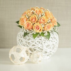An elegant floral display is captured in soft, natural light, showcasing a domed bouquet of approximately twenty to thirty peach and apricot roses, each flower arranged closely so that their velvety petals form a lush, rounded crown. The roses show gentle variations in colour, with deeper orange centres that fade to creamy, almost pastel edges, giving the arrangement a warm, sunset glow reminiscent of an early evening walk near Bentley Priory in Stanmore. Threaded between the rose heads are fine sprays of white baby's breath, their tiny blossoms creating a delicate mist that lightens the overall texture and adds a sense of romance, while glimpses of rich green leaves provide contrast and depth. The bouquet nestles securely in a large, spherical vase made from intricately woven white strips, forming an open lattice that feels modern yet organic, as if crafted by hand for a stylish home along Marsh Lane or around Stanmore Broadway. In the foreground, on a pristine white tabletop, two smaller decorative spheres rest casually: both are white and similarly woven, one with a neater, patterned structure and the other more irregular and artistic, subtly mirroring the main container. The background is a soft, neutral beige or greige wall with a fine, vertical texture that does not compete with the flowers, allowing every curve of petal and line of weaving to stand out clearly. The overall mood is calm and celebratory, ideal for a wedding reception, an anniversary dinner, or refined home styling, and feels like the kind of arrangement a local Stanmore florist would prepare with care for same-day delivery to nearby homes and venues.