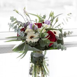 In the gentle mid-morning sunlight so typical of Stanmore's leafy suburbs, this bouquet embodies quiet refinement and warmth. At the center, velvety scarlet roses exude richness and emotional depth-their scent hinting at romantic walks through Canons Park. Crisp white gerberas, their petals like crepe silk, surround the roses, the contrast between cool whiteness and deep red highlighting an interplay of light and shadow. Arches of silvery eucalyptus leaves provide both a soothing backdrop and a touch of countryside tranquility, evoking the serene glades along Whitchurch Lane. Pale lilac lisianthus, some just beginning to open, add an air of anticipation and gentle beauty, much like the unfolding of spring in Stanmore's gardens. Fluffy white filler flowers add cloudlike volume and a playful softness, nestled between the bold blooms. The entire bouquet is anchored by lush, sculptural green foliage carefully wrapped around the base-a tacit reminder of the meticulous care Stanmore florists invest in every stem. The clarity of the glass vase allows a glimpse of verdant stems and cool water, further enhancing the sense of freshness and natural grace. This bouquet expresses heartfelt gratitude, quiet celebration, or sincere comfort, maturing into the perfect gesture for brightening a Stanmore home or marking life's treasured moments.