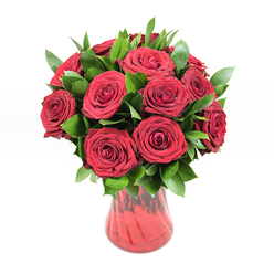 A lush, sculpted bouquet of approximately twelve to fifteen deep crimson red roses is showcased in a rich red glass vase, photographed against a clean white backdrop that allows every detail of the arrangement to stand out. The roses are at their prime, each bloom fully open and symmetrical, with tightly layered, velvety petals forming neat spirals that draw the eye inward. Their colour is a classic romantic red, intense and uniform, suggesting carefully selected premium stems rather than a mixed bunch. Between the blooms, a generous amount of dark green foliage, likely ruscus, provides structure and contrast. The leaves are small, pointed, and glossy, arranged so they peek out from the spaces between the roses, adding dimension and a sense of natural fullness, like something freshly arranged on a florist's bench just off Stanmore Broadway. The vase has a tall, gently curving profile, flaring slightly at the top and narrowing towards the base, with a subtle ribbed or swirling texture that catches the light and adds visual interest without competing with the flowers. Because the glass is translucent, the red appears to shift from deeper tones at the base to a lighter ruby glow near the rim, echoing the natural shadowing in the petals above. The crisp, even lighting creates soft highlights on the petals and leaves, while eliminating harsh shadows, so the bouquet appears timeless and serene. It's an arrangement that communicates commitment and passion, ideal for a surprise delivery to a home near Canons Park, a cosy dinner in Stanmore, or any occasion where classic red roses express more than words can manage.