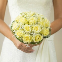Steeped in the enthusiasm of a Stanmore celebration, this bouquet radiates refined elegance beneath the soft indoor lighting of a wedding morning. Tightly clustered yellow roses, each blossom luminous and full, create a plush, rounded shape that echoes the joyful anticipation humming through Stanmore's leafy avenues. Sprigs of baby's breath, airy and iridescent, flow between the roses, adding a gentle contrast reminiscent of early dew in Canons Park. Gracefully coiled bands of deep green foliage or wire finish the bouquet at its base, lending both poise and structural beauty-a hallmark of Stanmore's talented florists. The bouquet is cradled lovingly in hands sporting delicate manicures and a single, meaningful ring, set against the subtly textured white of a wedding gown. The softened backdrop blurs away, drawing attention to the artistry at the center-a composition that captures the hope and serenity every Stanmore bride dreams of, making it the heart of any local wedding album.