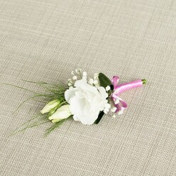 A refined wedding boutonniere is displayed on a softly woven, light beige fabric that looks like natural linen, the neutral background giving the composition a calm, timeless feel. At its heart is a fully open white lisianthus, its petals layered in delicate ruffles that curl back gracefully, creating a soft, cup-like shape. The flower appears freshly picked, with a luminous, almost velvety finish to the petals that suggests early morning preparation for a ceremony in Stanmore. To the left nestle two unopened lisianthus buds, long and slender, in creamy ivory flushed with pale green; they lean gently towards the main bloom, adding height, rhythm, and a sense of unfolding time. Threaded between them are fine sprigs of white baby's breath, their tiny star-like florets giving the piece a cloud-like lightness and an airy, romantic touch. Wispy blades of narrow, dark green foliage extend diagonally towards the lower left corner, giving the design movement, while one broader, richer green leaf sits behind the main flower, anchoring the composition like a subtle backdrop of garden foliage. At the base, the stems are tightly bound with a vivid fuchsia pink satin ribbon, elegantly tied into a small bow positioned near the bottom right. The ribbon's silky surface reflects the light, providing a bold yet tasteful contrast to the white flowers and muted background, like a splash of colour against a groom's suit at a Stanmore Hill wedding. The image is lit gently from above, showcasing the craftsmanship of a local florist and capturing the intimate, joyful mood of a special celebration, from church ceremonies to registry office vows nearby.