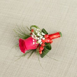 A carefully crafted boutonniere is displayed on a neutral, light beige woven fabric, the subtle texture of the background reminiscent of a linen suit jacket one might wear to a Stanmore wedding near Canons Park. At the centre, a single fuchsia pink rose rises proudly, its petals densely layered and slightly cupped, as though just beginning to unfurl into full bloom. The rose's colour is rich and saturated, a velvety pink with hints of magenta that immediately signals romance and joyful energy. Nestling at its base, tiny star-like blooms of white baby's breath form a soft halo, their airy presence creating a gentle transition between the vivid rose and the surrounding foliage. Intermixed with these delicate white flowers, small clusters of rounded, bright red hypericum berries lend a playful, almost jewel-like quality, their gloss catching the light and deepening the festive mood. Slender, feathery foliage, similar to asparagus fern, stretches outward in light green sprays, echoing the natural ease of nearby Bentley Priory Nature Reserve, while broader, darker leaves are artfully curled and layered to shape and support the design. The stems of the flowers and greens are tightly wrapped in a smooth, crimson satin ribbon, finished with a tidy bow that sits snugly at the base, making the buttonhole comfortable to wear and visually complete. The soft, overhead lighting casts faint shadows that emphasise each curve and contour, allowing the boutonniere's textures and colours to feel three-dimensional and alive-a small but meaningful floral accent perfect for grooms, ushers, or formal guests gathering around Stanmore's local churches and reception venues.