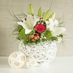 A stylish wedding-table arrangement is captured in this image, evoking the refined calm of a Stanmore dining room prepared for a celebration. The focal point is a spherical white vase crafted from interlaced strands, giving it an airy, open-weave pattern that feels both modern and organic, as if it were made from delicately bleached vines. Overflowing from this sculptural container is a considered mix of blooms. Tall white lilies rise above the rest, some fully open with broad petals arching outward to reveal gently speckled centres and golden stamens, others still tightly sheathed in green buds, adding vertical interest and a sense of unfolding time. Among these stately lilies sit rich red roses, their velvety petals tightly wrapped in perfect spirals; one main rose sits almost at the core of the piece, anchoring the design with an unmistakable symbol of love. Near the base, glowing green spherical flowers, likely green dianthus or button chrysanthemums, add a cushion-like texture and a burst of colour reminiscent of the lawns at Stanmore Golf Club under bright sunlight. Delicate sprays of white baby's breath are tucked between the larger blooms, forming little clouds of blossom that lighten the overall feel. Small, glossy red berries dot the arrangement, introducing a subtle nod to festive or winter occasions, making the piece as suitable for a Christmas wedding as for an elegant anniversary dinner. A variety of greenery, from fine, feathery strands to broader, darker leaves, fills out the silhouette, giving the design a lush, natural fullness without feeling heavy. The vase rests on a spotless white surface, and to the left, two woven decorative spheres in a soft beige tone echo the vase's pattern, balancing the composition. Behind everything, a neutral light-beige wall with gentle texture provides a calm backdrop, ensuring the crisp whites, deep reds, and fresh greens take centre stage for any special event in and around Stanmore.