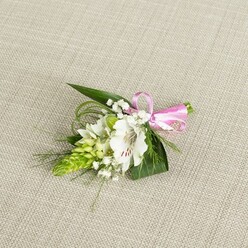 A compact floral boutonniere lies elegantly on a pale beige, linen-like cloth, the soft, woven texture creating a gentle backdrop that lets the floral work take centre stage. In the middle, a pristine white alstroemeria opens fully, its petals slightly ruffled at the edges and brushed with fine reddish-brown speckles that draw the eye down into the throat of the bloom. To the left, a striking column of fresh lime-green buds forms a tapered spike, each bud tightly nestled against the next, adding a bold, modern structure against the more romantic shapes around it. Wisps of white baby's breath weave through the arrangement, their countless tiny flowers giving a light, cloud-like softness that contrasts with the defined lines of the foliage. The greenery is thoughtfully layered: broad, glossy dark green leaves cradle the blossoms, while slimmer, grass-like strands arc outwards, adding lively movement and delicate texture. Tied neatly to the right of the stems is a bright pink satin ribbon, the bow small but perfectly formed, its sheen catching the light and bringing a playful yet refined accent to the otherwise white-and-green palette. Soft, even lighting bathes the piece, revealing the freshness of each petal and leaf, and suggesting flowers prepared that morning for a special event in Stanmore - perhaps a wedding near Stanmore Country Park or a prom at a local school. The overall feeling is one of poised calm and joyful expectation, the kind of finishing touch a local florist would hand to a guest just before they step out for an important celebration.