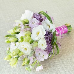 A radiant spring bouquet, crafted with expert care by a Stanmore florist, is lavishly arranged on a creamy, lightly textured linen that echoes the gentle hues of Stanmore's gardens. Lush white Lisianthus flowers bloom at the centre, their softly ruffled petals and warm yellow centres radiating elegance. Tender buds of lisianthus in pale white and lime green evoke freshness and new beginnings, while lavender-pink stock flowers add a soft, fragrant layer. Deep purple allium, boasting unique star-shaped florets, punctuates the arrangement, lending energy and structural beauty reminiscent of the sculpted landscapes around Stanmore Country Park. Curved blades of bear grass and textured green fillers introduce fluidity and natural movement, weaving through the bouquet as if following the gentle breeze across Stanmore's green expanses. The stems, gathered with precision, are wrapped in vibrant fuchsia satin and adorned with a coiled strand of classic white pearls-a harmonious balance of celebration and tradition. This bouquet, perfect for bridal parties or as inspiration for sophisticated events, embodies the joyful anticipation and serene atmosphere characteristic of Stanmore occasions, creating a stunning impression for any special gathering in this welcoming part of North London.