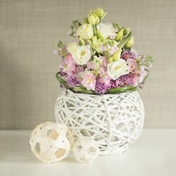A showcase of Stanmore's sophisticated approach to floral celebration, this arranged bouquet for wedding tables marries artful local craftsmanship with the timeless beauty of spring blooms. The centerpiece is abundant in airy lisianthus blossoms-creamy white and pale yellow-whose ruffled petals evoke morning sunlight streaming over Canons Park. These are lifted even further by slender stalks of blush and pale pink matthiola, granting height and gentle romance, while an undercurrent of lively purple filler flowers-suggestive of local asters or miniature alliums-brings vibrant depth. Enclosed in a contemporary spherical vase of open-work white rattan, each intricate strand reflected in the matching pair of decorative spheres to the side, the arrangement conveys tactile and visual delicacy. Deep green foliage forms a careful encircling border, reminiscent of Stanmore's green avenues. Resting atop a pure white table where soft, natural light highlights each textural nuance, the whole scene glows with calm invitation. A neutral, subtly textured beige backdrop elevates the bouquet's colours and warmth, making this a quintessential centrepiece for weddings, anniversaries, or elegant home gatherings in and around Stanmore-reflecting Flower Delivery Stanmore's skilled artistry.