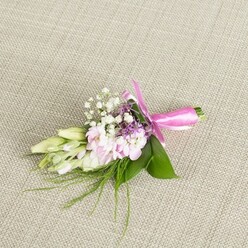 On a softly textured, pale beige linen backdrop, this elegant boutonniere or wrist corsage appears as if it has just been set down by a careful florist's hand, ready for a special Stanmore celebration. The background's fine weave and natural tone echo the look of a well-dressed table in a local venue near Stanmore Hill, allowing the colours of the flowers to glow without distraction. At the centre of the piece, a small cluster of multi-petalled soft pink blooms-resembling ruffled mini stocks or dainty garden blossoms-unfolds in layers, their petals shading gently from a deeper rose at the core to paler pink at the edges. Nestled throughout are airy tufts of white baby's breath, each tiny floret adding an almost mist-like softness and an old-fashioned charm. Here and there, discreet touches of lavender or deep purple flowers punctuate the pastel palette, providing depth and a refined, almost romantic contrast. Extending to one side, a line of creamy white to pale green buds, likely freesias or similar slender flowers still furled, introduces a sense of movement and anticipation, as though the buds may open slowly over the course of a wedding reception. The whole arrangement rests on a base of vibrant emerald leaves, smooth and broad, with fine, feathery sprigs of greenery reaching outward to suggest garden freshness. At the base, the stems are drawn tightly together and wrapped in a rich fuchsia satin ribbon, tied into an immaculate bow whose glossy loops and short trailing ends add a modern, celebratory accent. The gentle, even lighting captures every texture-from the cool satin to the velvety petals-inviting you to imagine this handmade detail worn on a lapel or wrist for a wedding, prom, or graduation around Stanmore and neighbouring Harrow Weald, where thoughtful floral touches elevate the entire day.