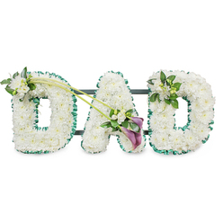 The image presents a graceful "DAD" funeral tribute, a floral arrangement that could easily be pictured resting beside a coffin in a Stanmore chapel or at a graveside overlooking the greenery of Canons Park. Each of the three letters is constructed as a sturdy, upright block, closely covered with snow-white chrysanthemums or similar small button blooms. The flowers are massed tightly together, creating a plush, uniform texture that looks soft to the touch and radiates purity and calm. Around the outer edges of all three letters runs a ruffled band of emerald green satin ribbon, meticulously pinned to follow every curve, providing a crisp frame that highlights the contours and adds a fresh, foliage-like border. Faint glimpses of a dark metal frame can be seen behind the letters, indicating the tribute is designed to stand proudly in a row during a funeral, wake, or memorial service in Stanmore.   

Subtle yet expressive floral embellishments bring personality and emotion to the piece. On the upper left side of the first "D", a small spray of white freesia and vibrant green foliage is gently tucked into the chrysanthemums, the freesias offering a lighter, more open texture and suggesting a discreet, sweet fragrance. From this accent, two slender green stems extend diagonally across toward the central "A", linking the letters and creating a sense of flow. At the point where they meet the "A", an asymmetrical focal spray draws the eye: two deep purple calla lilies, with their sculpted trumpet-shaped blooms, are nestled among additional white freesia flowers and glossy leaves. The rich purple hue provides a poignant, regal contrast, underscoring the solemnity of the tribute. On the top right curve of the final "D", a smaller, coordinating cluster of white blooms and foliage mirrors the first, bringing visual harmony. The overall impression is one of quiet strength, love, and respect for a father, conveying the heartfelt sentiments of family and friends across Stanmore.