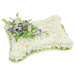 A meticulously composed funeral pillow, formed from hundreds of small, immaculate white chrysanthemum heads that create a plush, uniform surface reminiscent of a sleeping cushion. The floral rectangle is softly rounded at the corners and slightly raised in the centre, giving it a comforting, pillow-like silhouette. Along its perimeter a pale lime-green satin ribbon has been pleated into an even, scalloped border, the cool green lending a gentle, hopeful contrast to the sea of white. In the top-left corner a considered accent spray adds quiet colour: layers of white lisianthus sit beside rich purple lisianthus, their ruffled petals and emerging buds offering both contrast and tenderness. Wisps of feathery fern and slender foliage are threaded through the cluster, while a darker green leaf wrap neatly conceals the stems beneath, keeping the bouquet refined and dignified. The overall effect is one of serene texture - a soft visual hush where the chrysanthemums' round faces and the lisianthus' layered cups speak of purity and remembrance. The image is lit with soft, even light against a white backdrop and a subtle shadow beneath, suggesting the arrangement is ready for placement at a chapel service or graveside in the Stanmore area near Stanmore Broadway or Canons Park. Prepared by Flower Delivery Stanmore with experienced hands, this tribute balances formality and comfort: white for peace, purple for respect, and green for gentle renewal, sent with care to console those gathering to remember.