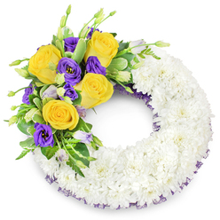 This circular funeral wreath is composed with careful, calming intent: a dense ring of pristine white spray chrysanthemums forms the majority of the piece, their tiny petals layered so closely that the surface reads like a soft, luminous cloud. The wreath's open centre keeps the classic 'O' silhouette intact, while a concentrated floral spray placed on the upper left provides a poignant focal point. Bright yellow roses, fully unfurled and warm as late-afternoon light, sit at the forefront of that spray, their gentle scent hinted at beneath the chrysanthemums' quiet presence. Intermingled are deep purple lisianthus, their satin-like petals and ruffled edges creating a rich counterpoint; a few unopened buds in paler lavender and green add freshness and natural rhythm. Sprigs of glossy green foliage - including rounded, slightly variegated leaves and fine ferny stems - weave through the cluster, offering structural support and visual relief between the colours. A slender band of dark purple mesh or crinoline is visible along the inner and outer rims, a discreet finishing touch that references the lisianthus tone and adds subtle continuity. The studio light is soft and even, accentuating the texture of the chrysanthemums and the velvety depth of the roses and lisianthus without dramatic shadow. The overall impression is dignified and comforting, an arrangement intended to express sympathy, lasting remembrance and gentle consolation for funeral or memorial settings in the Stanmore area and neighbouring Harrow.