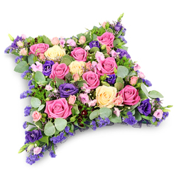 This image captures a beautifully detailed cushion-shaped funeral tribute, composed entirely of flowers and foliage so that it resembles a soft, floral pillow at rest. The dominant blooms are roses, their rounded heads and spiralling petals showcased in shades that range from deep, rich fuchsia to gentle blush pink and creamy pale yellow. Many of the roses are fully open, revealing intricate centres and velvety texture, while a few partially open buds introduce a feeling of life still unfolding. Between these roses weave stems of royal-purple lisianthus, their layered, ruffled petals adding depth, shadow, and a rich note of contemplation to the colour harmony. Small clusters of lavender-toned filler flowers, likely statice or limonium, appear throughout the design, filling every gap so the eye meets only petals and leaves, never bare foam or stems. Around the edges and peeking between the flowers, a selection of greenery creates a soft frame: rounded, silvery eucalyptus leaves with their subtle blue-green cast, and finer, fern-like foliage that adds delicate feathery lines. The overall shape is a gently domed rectangle with rounded corners, evoking comfort and the idea of placing a head on a pillow for a final, peaceful sleep. Set against a white background, the tribute's colours seem especially clear and serene. It feels like a piece that might be delivered to a quiet service in Stanmore, perhaps at a local church off Church Road or near Canon's Park, representing a family's love and respect. The arrangement speaks of tenderness, gratitude, and remembrance, providing a graceful, floral voice when spoken words can be hard to find.