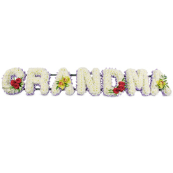 A long, horizontal floral tribute spells out the word GRANDMA in bold capitals, captured against a bright, uncluttered white background that allows the detail of the design to shine through clearly. Each individual letter is filled with tightly packed white chrysanthemums, layered so generously that they form a soft, padded surface, evoking comfort and serenity. Around the bottom and outer edges of the letters runs a fine trim of pale lavender flowers, lending a cool lilac halo that carefully defines the contours and adds depth without overpowering the purity of the white. Carefully chosen accent blooms punctuate the tribute, like small chapters in a family story. On the lower section of the initial G, three deep red roses, lush and velvety, are gathered together with dark green foliage, their rich colour suggesting heartfelt affection and remembrance. The first A showcases two yellow-green orchids perched neatly on its crossbar, each blossom with a delicately speckled reddish-brown centre. The following letters R, N, and D are left unadorned, their simplicity giving a feeling of calm respect. On the lower right stroke of the M, another trio of red roses echoes the floral cluster at the start, and the final A repeats the orchid detail with two more chartreuse blooms resting among the white petals. The letters are attached to a slim, dark metal bar along the back, ensuring stability when the tribute is displayed at a Stanmore funeral service, whether at a local church near Stanmore Broadway or at a crematorium reached via the leafy roads around Old Church Lane. The arrangement conveys a quiet, composed elegance, ideal for honouring a cherished grandmother with grace.