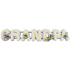In this image, the word GRANDPA is formed by a series of large floral letters, arranged horizontally and floating against a clean white background, echoing the tranquil atmosphere of a Stanmore funeral service. Every letter is shaped with precision, then densely covered with small white blooms, likely chrysanthemums or carnations, so that the flowers form a seamless field of gently ruffled petals. The texture is soft and inviting, reminiscent of a white blanket of blossoms laid out in quiet tribute. The dominance of white conveys innocence, gratitude, and the serenity that often surrounds a final farewell. Threaded into this gentle expanse are carefully placed clusters of rich purple and lilac flowers, framed by sprigs of glossy green foliage. These accents sit within the curves and angles of the letters-the hollow of the G, the triangular pockets of the As, and the rounded spaces of the D and P-creating subtle, jewel-like highlights without overwhelming the peaceful design. A dark green structural bar runs along the base of the letters, almost entirely hidden by the foliage and petals, providing strength and stability for display at a church, crematorium, or graveside around Stanmore or nearby Edgware. The arrangement feels like a lovingly considered choice for families who want a clear, heartfelt message of farewell for a grandfather, something that will read beautifully in photographs and in person as mourners pause to reflect, perhaps after a slow walk up from Stanmore station or along the quiet streets leading to the service. It's a tribute that balances craftsmanship with emotion, allowing flowers to speak the words that are sometimes hard to say aloud.