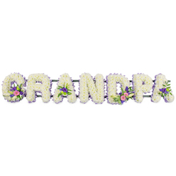 The image portrays a beautifully balanced funeral flower tribute spelling out the word GRANDPA, laid out horizontally and isolated against a bright white background, as if waiting to be placed beside a coffin in a Stanmore chapel or synagogue. Each letter is solid and full, built entirely from tightly clustered white chrysanthemums whose small, multi-layered petals give a soft, velvety depth, inviting thoughts of gentle hugs and shared stories with a cherished grandparent. Around the edge of every letter runs a fine outline of delicate lilac and purple florets that crisply trace the contours, ensuring each character stands out clearly while adding a calming wash of colour. At thoughtfully chosen points, accent sprays bring the tribute to life: the G and first A display small bouquets of white lily-like blooms framed by bright pink or fuchsia flowers and deeper purple accents, with glossy greenery peeping through. The D and another A mirror this look, including occasional bright green button chrysanthemums that offer a fresh, lively note without disturbing the solemn mood. A slim dark green bar is visible behind the letters, showing how the tribute is quietly supported so that GRANDPA remains perfectly aligned and readable from a distance. The lighting is even and soft, carving gentle shadows that emphasise the three-dimensional shape of each letter and the meticulous work of the florist. This arrangement feels personal and heartfelt, the sort of bespoke condolence tribute families in Stanmore might choose for a funeral or graveside service, conveying deep love, respect, and gratitude for a grandfather's life and legacy through the language of flowers.