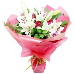Set against a clean white background, this bouquet looks like it has just been carefully arranged in a Stanmore florist's studio, capturing that moment when everything comes together in perfect balance. The focal point is a cluster of white Oriental lilies, their large, trumpet-shaped blooms opening outwards with a quiet sense of drama. Each lily has smooth, slightly recurved petals that create a star-like form, drawing the eye towards the centre where long stamens with dark brown, almost purplish anthers stand in sharp contrast to the pure white. Between these open flowers, several elongated buds in shades of green and cream remain tightly closed, promising that the bouquet will continue to develop and change over time. Threaded through and beneath the lilies are deep red roses, many of them in bud, their short stems carrying compact, velvety heads that add an intimate, romantic undertone. Together, these flowers rest on a foundation of lush green foliage: broad, glossy leaves provide depth and structure, while slim, grass-like blades arc upwards and outwards, suggesting gentle movement like the breeze you might feel walking through Stanmore Country Park. The entire design is wrapped in voluminous, glossy bright pink cellophane that has been gathered into generous, crinkling layers around the stems. The semi-translucent wrapping allows hints of dark green and shadow to show through, giving the composition a sense of dimension and softness. At the base, a narrow lime green ribbon is tied with precision, adding a fresh, contemporary twist that stops the pink from feeling overly sweet. The result is an arrangement that feels celebratory yet sophisticated, ideal for same-day delivery in Stanmore as a romantic surprise, a thoughtful thank-you, or a joyful gesture for birthdays and anniversaries.