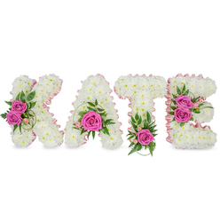 This personalised floral name tribute reads KATE in substantial, freestanding letters, each letter lovingly constructed from a dense mass of small, pure-white chrysanthemums that give the composition a pillowy, cloud-like texture. A pale-pink satin ribbon outlines the inner shapes of the K, A, T and E, softening the block letters with a gentle, feminine line that gleams faintly in soft daylight. Bright hot-pink roses act as emotional punctuation: on the K two fully opened roses sit to the left, cradled by mixed green foliage; the A bears one bold rose low in its curve with delicate pink bud accents and fine trailing greenery; the T is finished with a single vivid rose at its base surrounded by fern-like fronds; and the E is decorated with a cluster of two roses on its right side, balanced by variegated leaves and small pink fillers. The chrysanthemums are tightly packed, their tiny petals forming a uniform, tactile surface that suggests careful handwork and professional structure. Light falls gently across the top surfaces, creating soft shadows that reinforce the three-dimensionality of the piece and its freestanding presence. A subtle floral perfume and the fresh, leafy scent of greenery lend an olfactory softness to the visual calm. Designed with sympathy and dignity in mind, this tribute would be fitting for a memorial gathering in Stanmore - whether delivered to a chapel near Bentley Priory or placed beside a service on Stanmore Hill - reflecting thoughtful craftsmanship and a quiet sense of love.