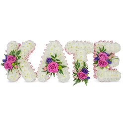 A lovingly composed memorial arrangement spells the name KATE in four upright, three-dimensional letters, each formed from thousands of compact white chrysanthemums that create a uniformly plush, cloud-like surface. The soft studio lighting highlights the subtle layers of petals and the pale blush of a thin pink satin ribbon that delicately edges every contour - an artisan touch that adds gentle definition without overpowering the purity of the white blooms. The tactile contrast between crisp chrysanthemum heads and the smooth sheen of ribbon gives the letters an intimate, handcrafted presence.

Strategically placed accent clusters bring warmth and colour: large fuchsia-pink roses provide rich, velvety focal points; smaller pastel pink spray roses offer tenderness and gradation; and fine sprays of deep purple or blue flowers, likely statice or compact lisianthus, introduce cool, measured contrast. These bouquets are cushioned and carried by varied greenery - ferny fronds, rounded eucalyptus leaves, and glossy foliage - which together lend texture, scent, and an organic silhouette. Floral placement is intentional: a striking cluster on the K's diagonal, a gentle grouping at the A's lower right, a base cluster on the T, and two balanced groupings on the E's middle and bottom bars.

Prepared with the care of a local Stanmore workshop, this bespoke tribute evokes solace and remembrance. Flower Delivery Stanmore can create similar personalised name letters for funerals, wakes, or memorials across Stanmore and nearby Canons Park, offering a quiet, expressive way to honour a loved one.