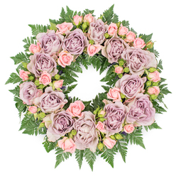An overhead view reveals a perfectly rounded floral wreath arranged on a pure white surface, its circular form symbolising unbroken love and remembrance. The design is dominated by roses in refined, muted colours, creating a calm, contemplative mood. Large, open roses in dusty lavender and antique mauve are placed with measured regularity, their many-layered petals forming soft spirals that draw the gaze inward. These blooms have a velvety texture that suggests both tenderness and quiet strength. Threaded between them are clusters of blush pink spray roses, their smaller size and varied stages of blooming adding delicacy and nuance to the composition. Some of these petite roses are still in tight bud, hinting at stories that remain tucked away in the hearts of those who grieve, while others are just beginning to open, echoing the way cherished memories gradually unfold over time. A substantial foundation of dark green fern foliage underpins the entire wreath, each frond's serrated leaflets overlapping to create a rich, textural frame. The vibrant green offers a grounding contrast to the soft purples and pinks, not unlike the comforting green spaces around Stanmore, from peaceful residential gardens to nearby parks where people gather to remember and reflect. Prepared with the attentiveness of an experienced local florist, this wreath feels suited to a funeral or memorial service at a Stanmore church, crematorium, or cemetery. The soft, even lighting and absence of visual clutter focus all attention on the flowers themselves, making the wreath appear both elegant and sincere, a floral expression of sympathy, respect, and enduring affection.
