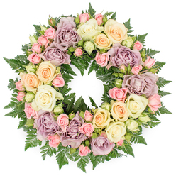 A full, round memorial wreath made almost entirely of roses emerges clearly against a bright white background, every bloom arranged to create an even, flowing circle of remembrance. The colour palette is soft and muted, with creamy white and ivory roses mingling with gentle peach, pale yellow, and blush pink blooms, then deepening into more unusual dusty lavender and mauve tones that add a subtle touch of character and distinction. Large, open roses form the main structure of the design, their layered petals opening like quiet, unfolding stories, while clusters of smaller spray roses and tight buds slip between them, creating intricate patterns and a sense of continuity. A bed of deep green fern fronds cradles the flowers, the slender, divided leaves peering out between petals and around the outside edge, giving the wreath body, contrast, and a hint of the natural greenery around Stanmore's parks and gardens. The circular shape is perfectly even, with no dominant side, symbolising an endless loop of love, memory, and connection. Imagined resting at a memorial service in a Stanmore chapel or laid by a headstone overlooking Bentley Priory Nature Reserve, this wreath feels composed, calm, and deeply respectful. Light in the image falls softly, enhancing the gentle gradations of colour and the satin-like texture of the petals. As an experienced local florist, Flower Delivery Stanmore might create such a piece for families seeking a traditional yet personal tribute, one that speaks in quiet tones of condolence, gratitude, and the wish to honour a life with beauty and care.